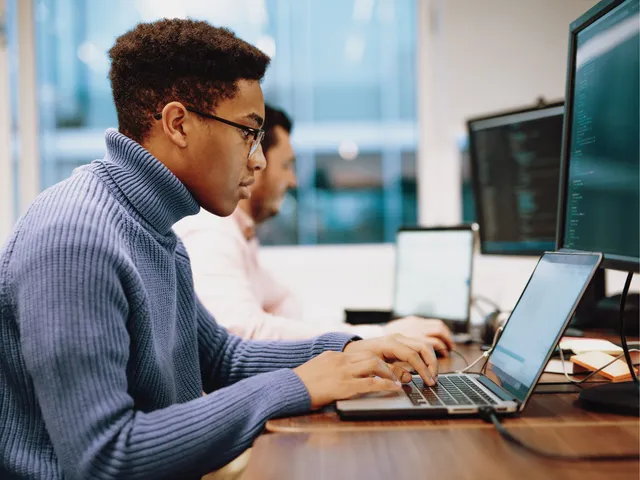 Focused professionals working at laptops and desktop monitors in a modern office, analyzing code or technical data.