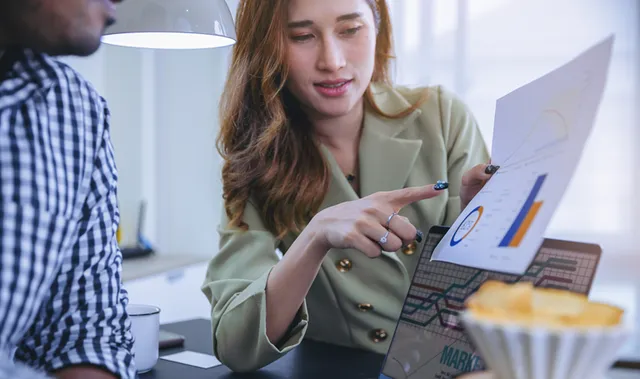 Woman in a green blazer pointing at a laptop screen showing analytics charts while explaining data to a colleague.
