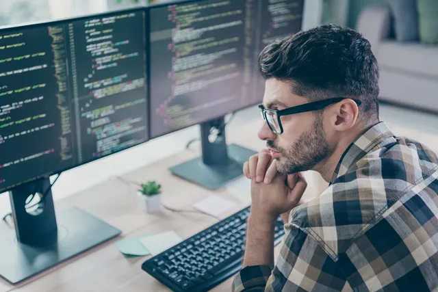 A person in a plaid flannel shirt and glasses rests their chin on their hand while reading code displayed across two large monitors at a bright home workstation.