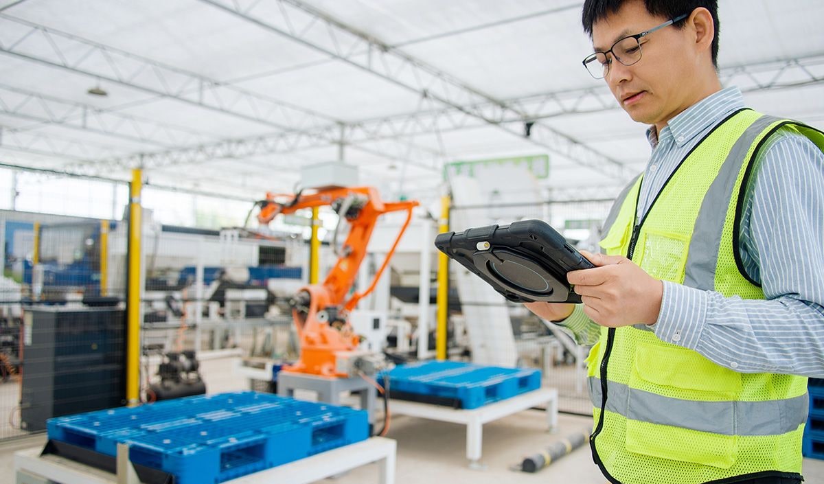 Worker in a safety vest using a tablet on a modern automated manufacturing factory floor with a robotic arm in the background.