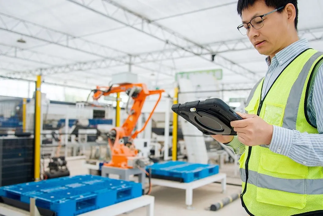 Worker in a safety vest using a tablet on a modern automated manufacturing factory floor with a robotic arm in the background.