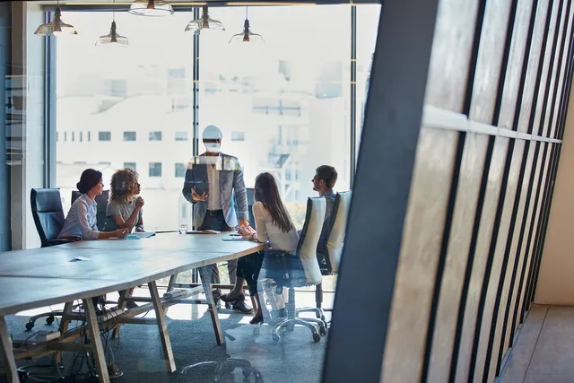 A standing presenter leads a meeting with four seated colleagues at a long table in a bright boardroom with floor-to-ceiling windows showing a city beyond.