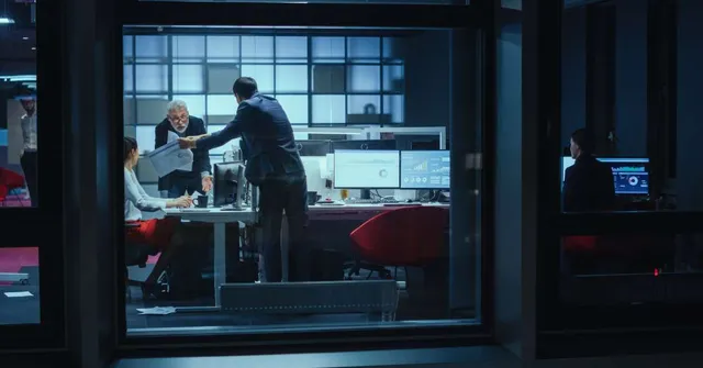 Modern office at night with professionals working, including a handshake in the foreground and data analytics dashboards on monitors.