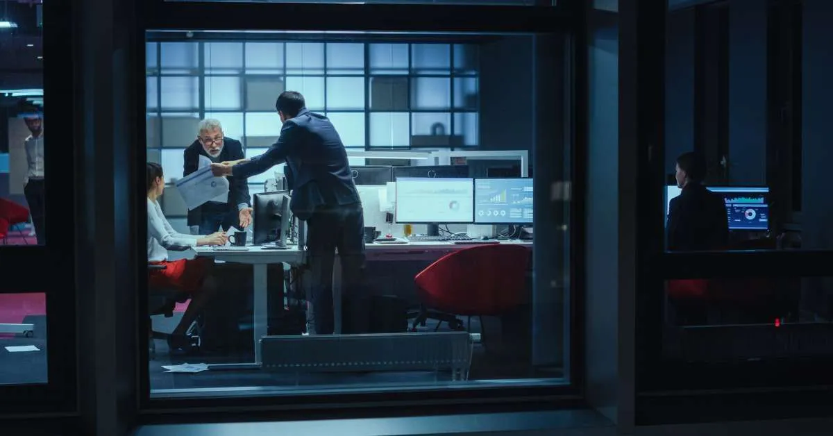 Modern office at night with professionals working, including a handshake in the foreground and data analytics dashboards on monitors.
