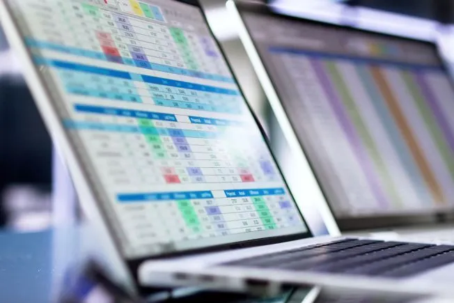 Two laptops side-by-side in an office showing data tables of company financials.