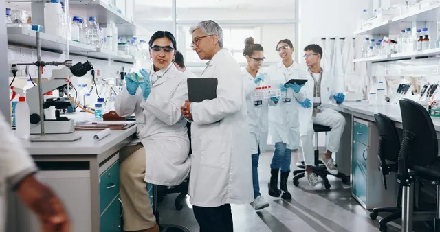 A team of scientists in lab coats and safety glasses examine a petri dish, a tablet, and test tubes in a bright research laboratory.