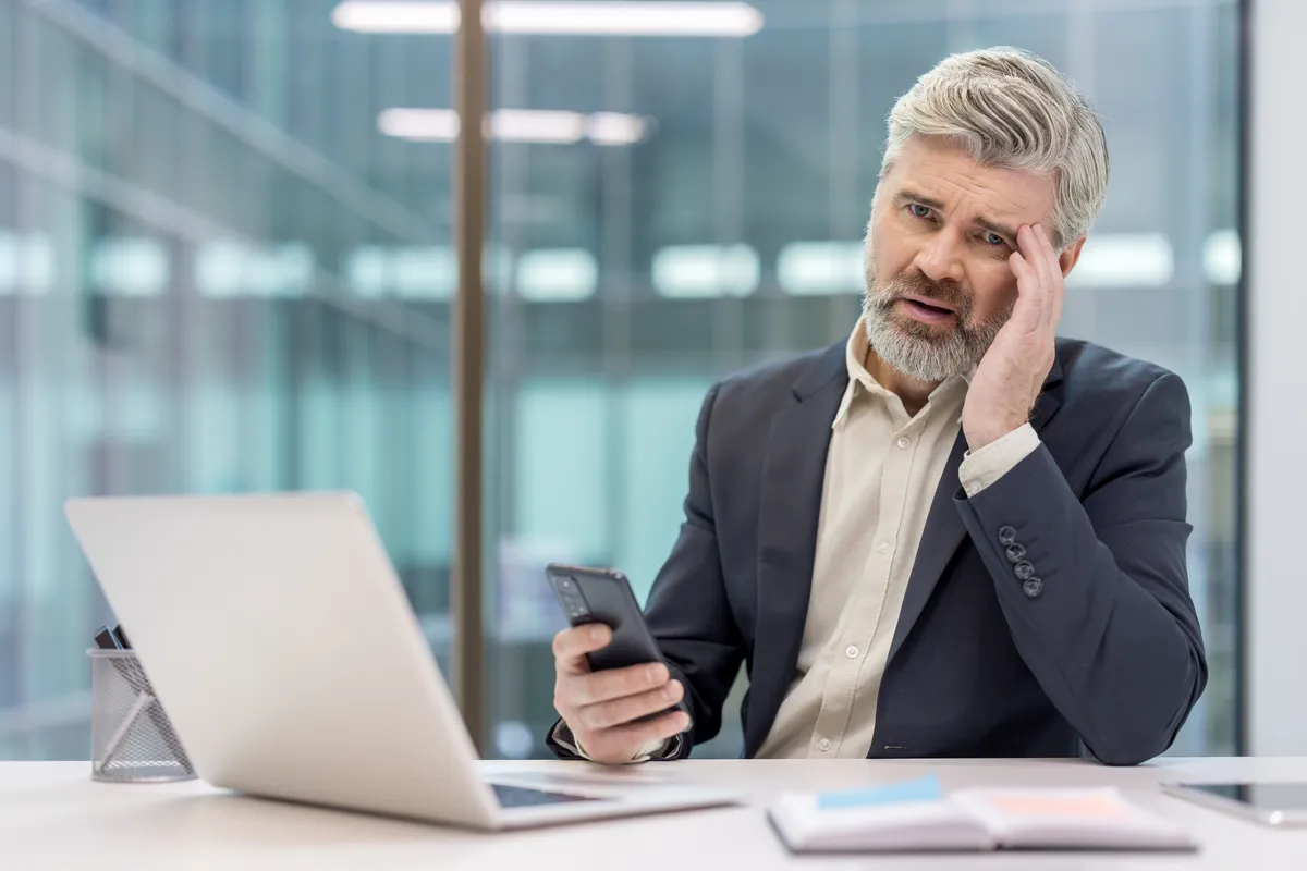 A person with a gray beard sits at a white desk with an open laptop in a glass-walled office, looking at a phone.