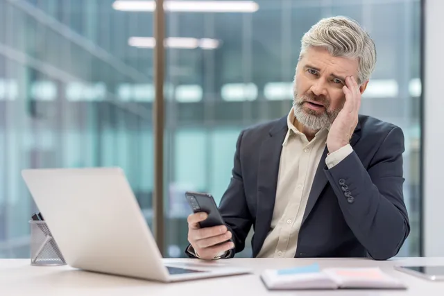 A person with a gray beard sits at a white desk with an open laptop in a glass-walled office, looking at a phone.