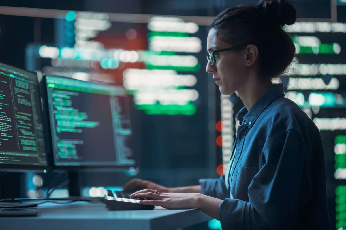 Person in glasses and a blue shirt working at a computer in a data center, with monitors displaying green code and data visualizations.