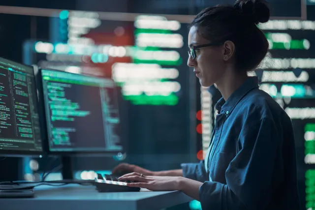 Person in glasses and a blue shirt working at a computer in a data center, with monitors displaying green code and data visualizations.