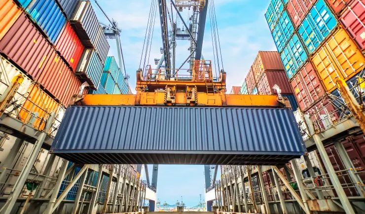 Yellow crane lifting a dark blue cargo container at a busy international shipping port terminal.