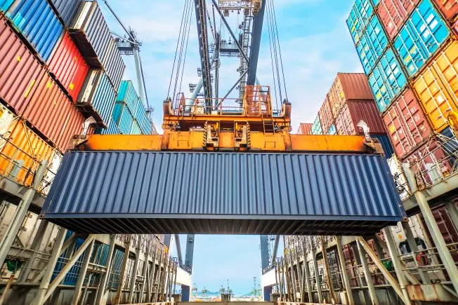 Yellow crane lifting a dark blue cargo container at a busy international shipping port terminal.