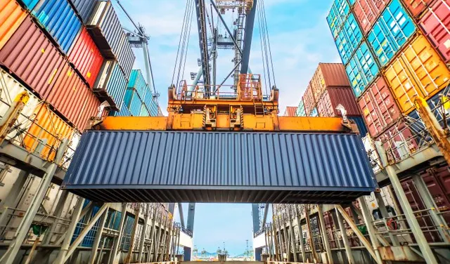 Yellow crane lifting a dark blue cargo container at a busy international shipping port terminal.