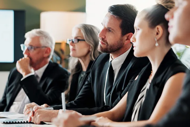 Five people in dark business attire sit in a row at a conference table, looking toward a screen displaying charts.