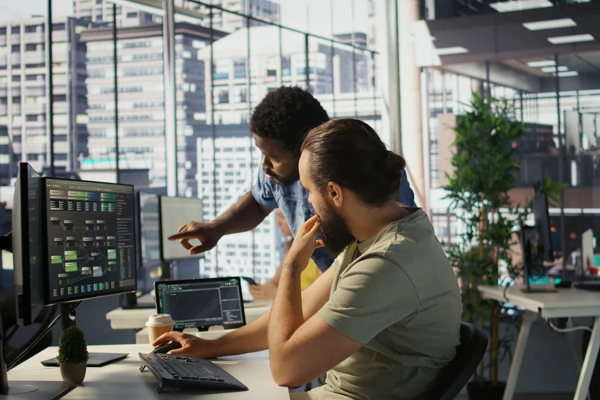 Two colleagues review financial charts and training software data on dual monitors at a desk in a high-rise office overlooking a sunlit city skyline.
