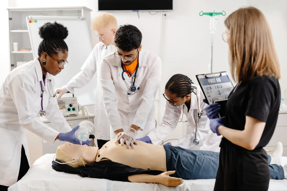 Four medical students in lab coats practice CPR and bag-mask ventilation on a training mannequin while another student holds a first-aid clipboard.