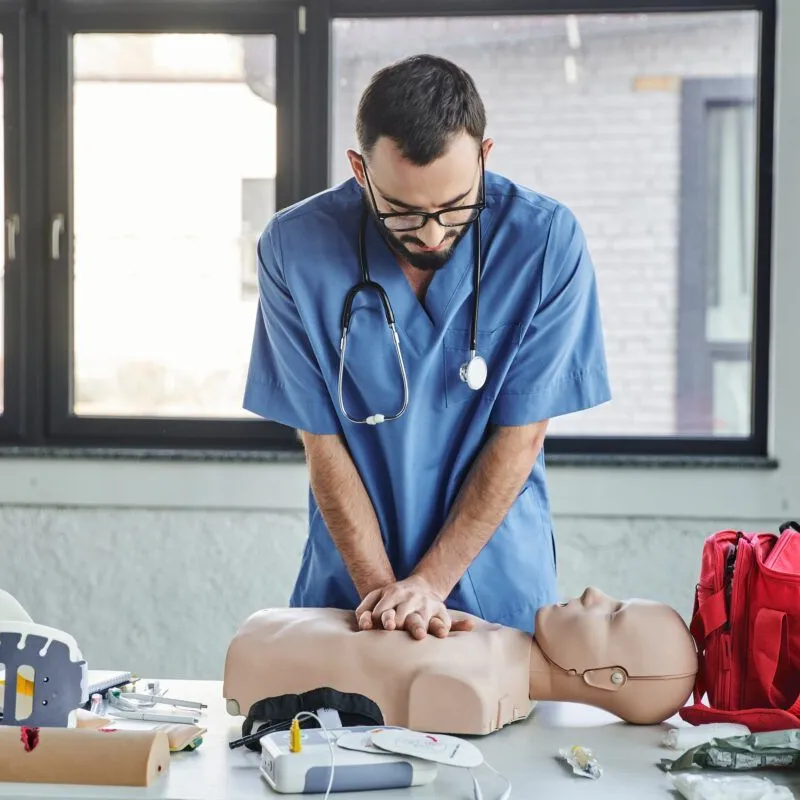 Young paramedic in blue uniform and eyeglasses practicing chest compressions on CPR manikin near defibrillator and first aid kit during medical seminar, life-saving skills development.
