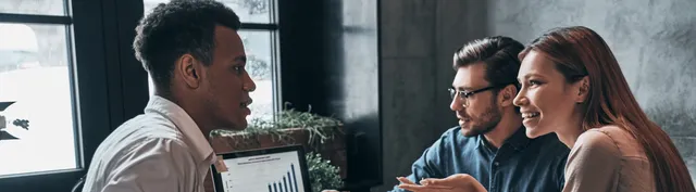 Three professionals in a modern office, one holding a tablet with a bar chart, engaged in a casual business discussion.