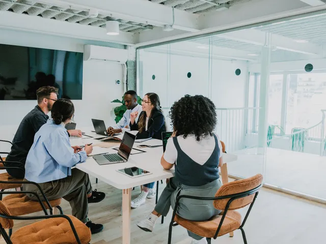 Diverse team of five professionals collaborating around a table with laptops in a modern office with floor-to-ceiling windows.
