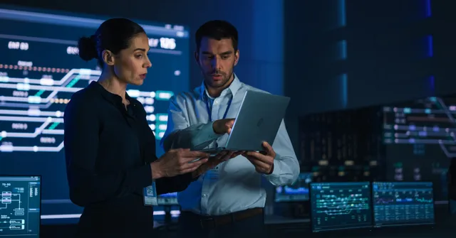 Two professionals reviewing a tablet in a high-tech control room with blue digital monitoring displays in the background.