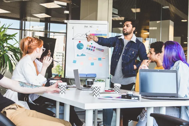 A standing presenter in a denim jacket points to colorful data charts on a flipchart while four colleagues with laptops and coffee mugs watch from a meeting table.