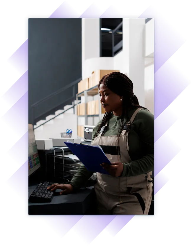Woman in work overalls using a desktop computer while holding a clipboard in a stockroom.