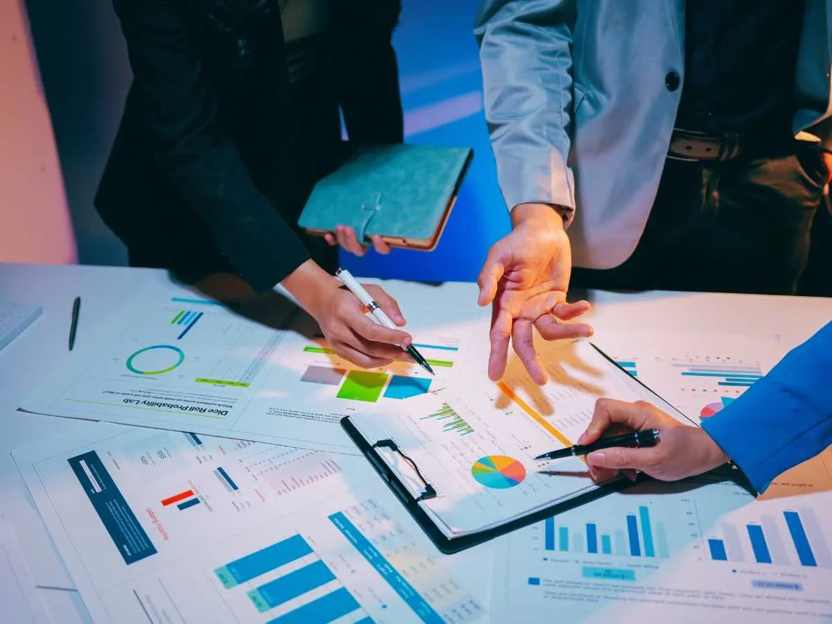 Three people's hands point at and highlight various colorful bar charts, pie graphs, and data reports spread across a desk, illustrating a collaborative team review of performance or compliance metrics.