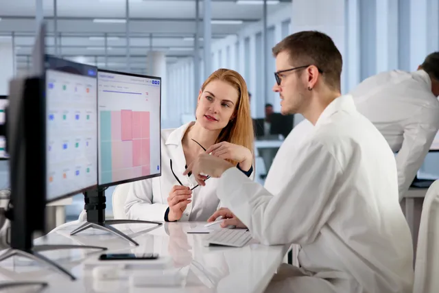 Two people in white lab coats sit at a workstation reviewing charts on large monitors in a bright, modern open-plan office.