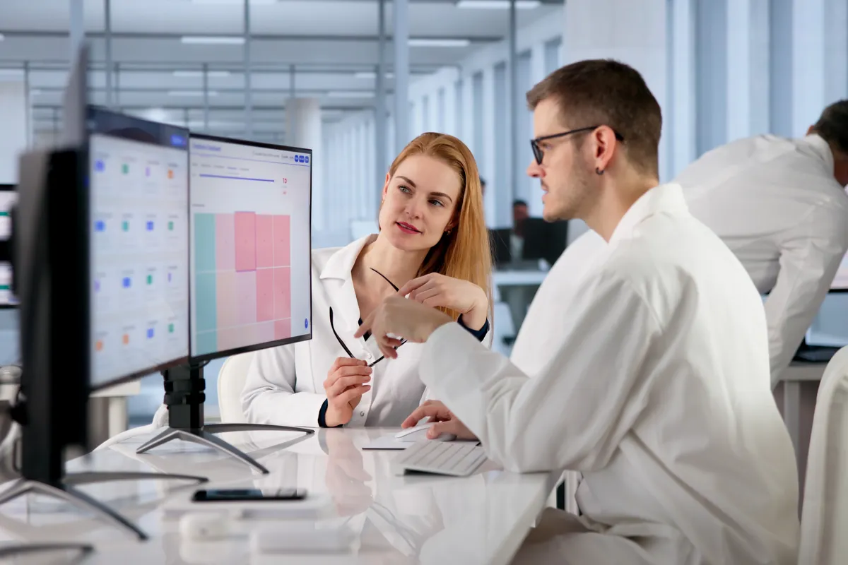 Two people in white lab coats sit at a workstation reviewing charts on large monitors in a bright, modern open-plan office.