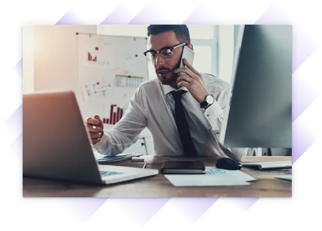 Very busy professional on a phone call while working on a laptop at a desk with charts and monitors.
