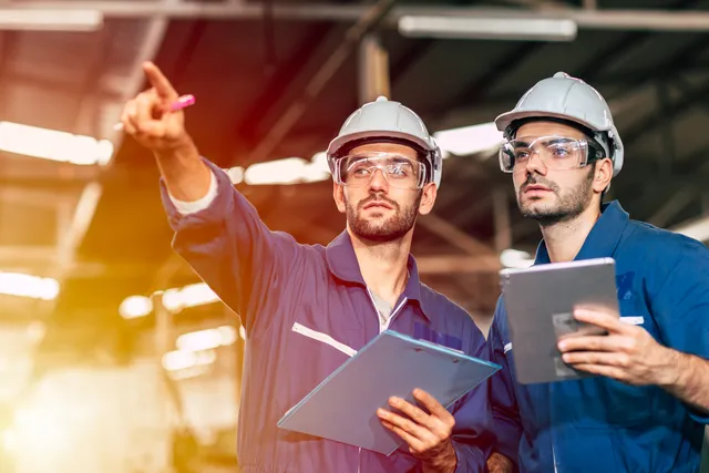 Two workers in blue coveralls, white hard hats, and safety glasses review a clipboard and tablet inside an industrial facility while one points upward.