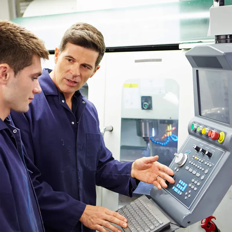 Two male technicians in dark blue uniforms operating a CNC machine control panel in an industrial manufacturing facility.