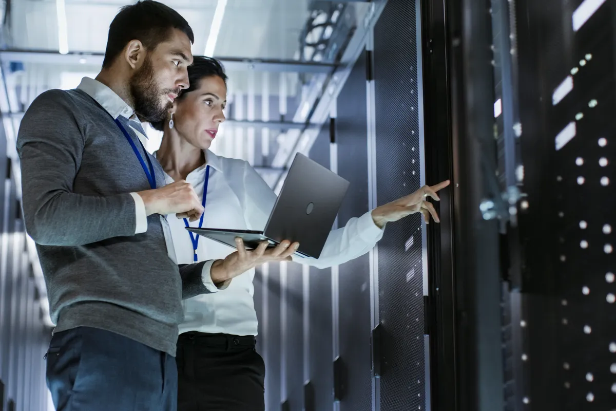 Two IT professionals stand together in a data center aisle, holding an open laptop and pointing toward a row of illuminated server racks.
