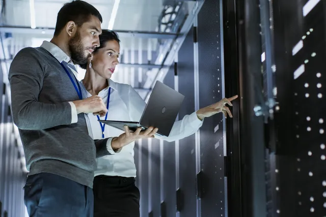 Two IT professionals stand together in a data center aisle, holding an open laptop and pointing toward a row of illuminated server racks.