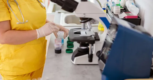 Healthcare worker in a yellow uniform handling laboratory sample containers at a clinical workstation.