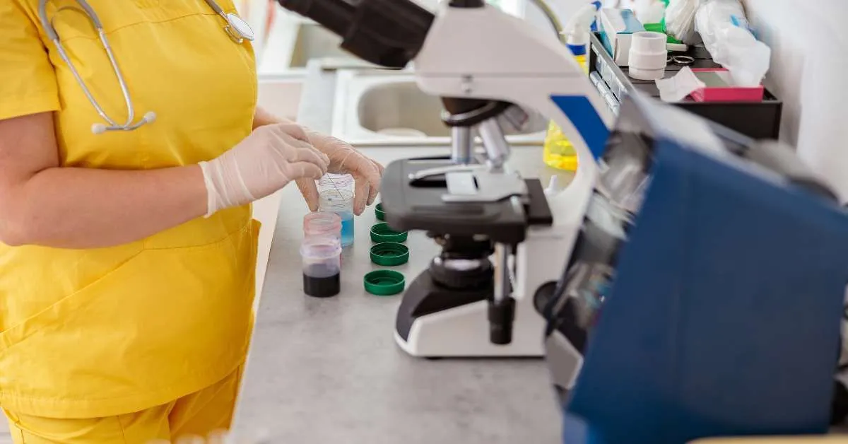 Healthcare worker in a yellow uniform handling laboratory sample containers at a clinical workstation.