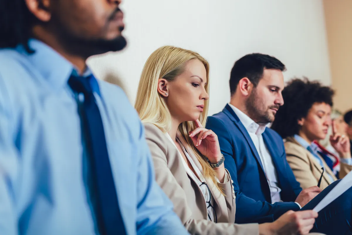 A row of people in business attire sit along a plain office wall, one resting a hand on her chin while another reviews papers.