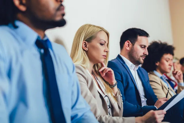 A row of people in business attire sit along a plain office wall, one resting a hand on her chin while another reviews papers.