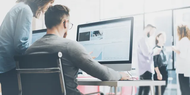 Man and woman reviewing a desktop monitor displaying a data analytics dashboard in a bright modern office.