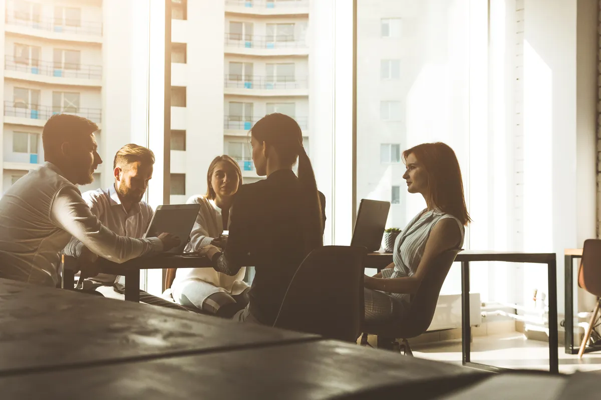 A small team gathers around laptops at a long conference table in front of floor-to-ceiling windows with backlit city buildings outside.
