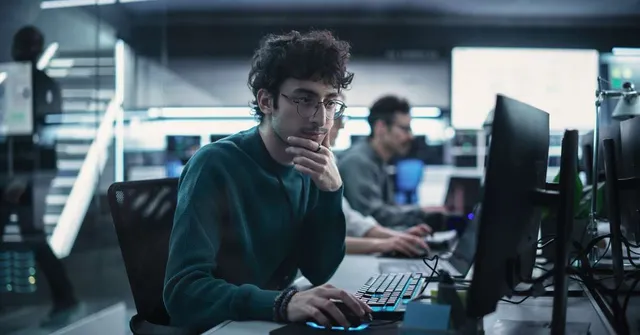 Young man with glasses working at a computer in a busy open-plan tech office with multiple monitors and neon lighting.