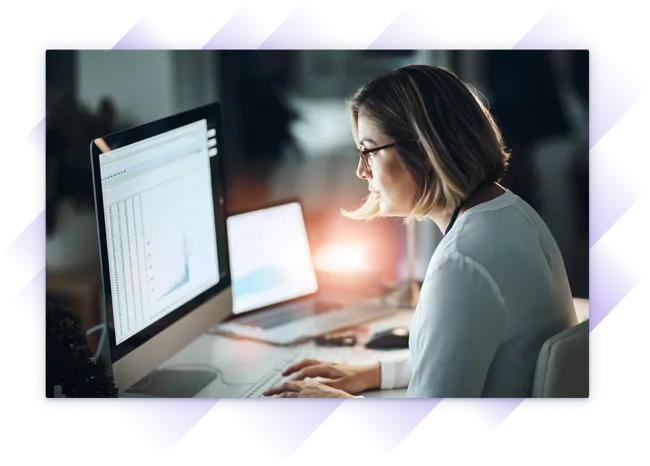 Woman looking closely at two monitors as the sun goes down.