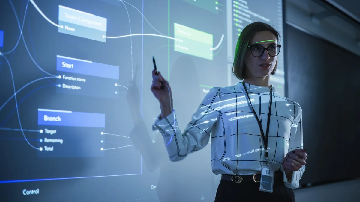 A presenter holding a stylus stands in front of a glass wall projecting a software flowchart and code, surrounded by data visualizations.