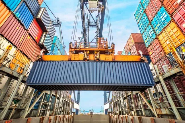 A crane lifts a large blue shipping container above stacked rows of colorful containers at a busy port under a partly cloudy sky.