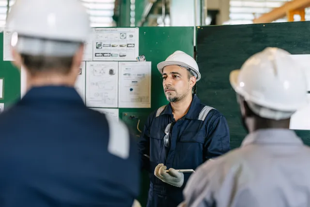 A worker in a white hard hat and navy coveralls holds a tablet beside a control panel and instructional diagrams, addressing two colleagues in hard hats inside an industrial facility.
