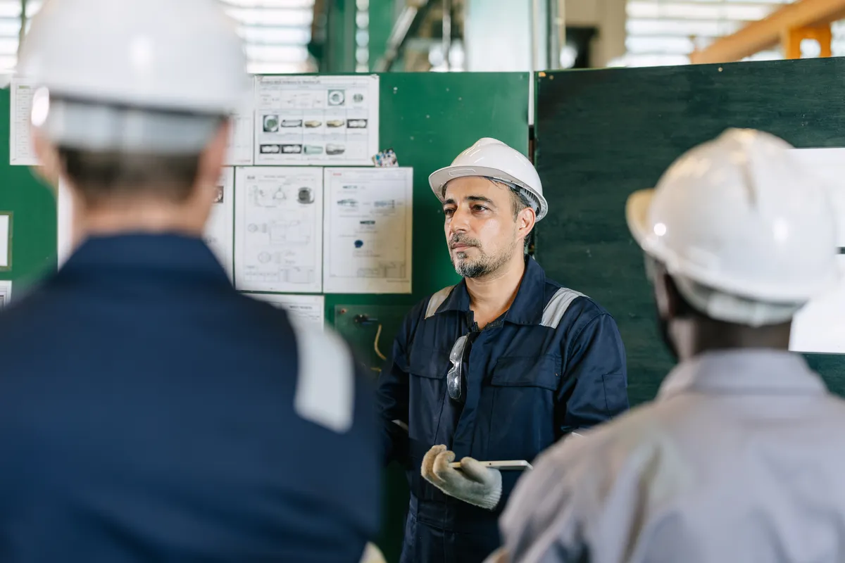 A worker in a white hard hat and navy coveralls holds a tablet beside a control panel and instructional diagrams, addressing two colleagues in hard hats inside an industrial facility.