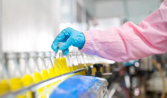 Laboratory worker in a pink lab coat and blue gloves holding a glass vial of yellow liquid in a pharmaceutical manufacturing facility.