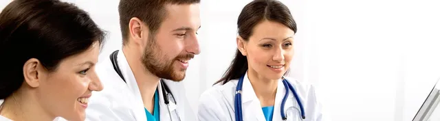 Three people in white medical coats with stethoscopes smile while reviewing information together on a screen out of frame.
