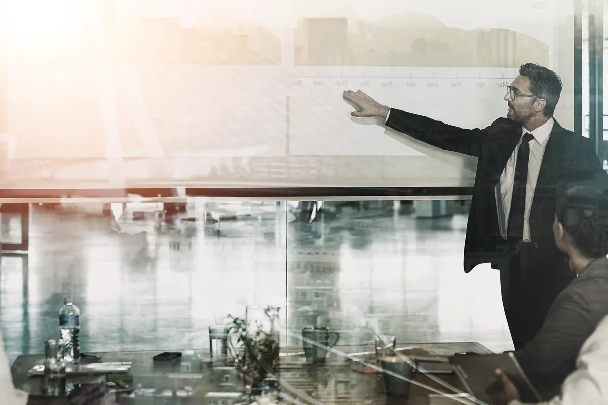 A presenter in a dark suit gestures toward a screen during a boardroom meeting in a wide double-exposure composite layered over a city waterfront skyline.