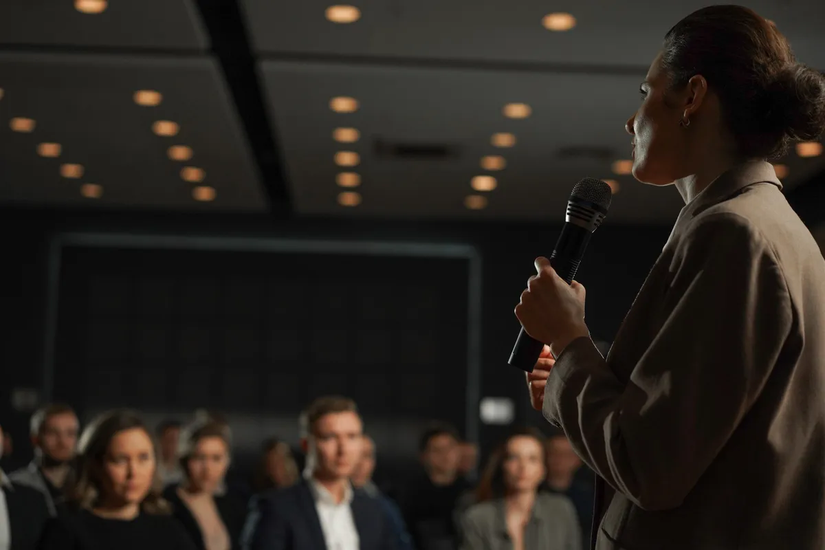 A woman in a tan blazer holds a microphone and addresses an audience in a dimly lit conference hall, with warm ceiling lights overhead and attendees seated in soft focus in the foreground.